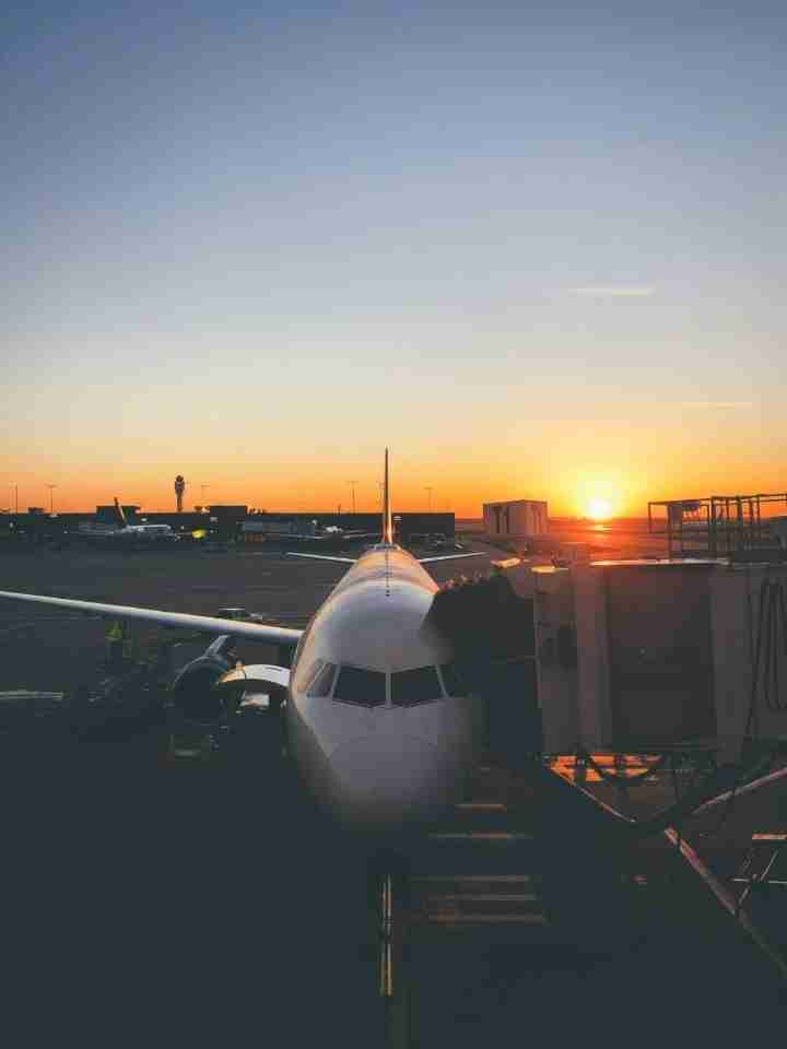 Front view of a passenger jet at a gate during sunset, with a jet bridge on the right and warm orange sky on the horizon.